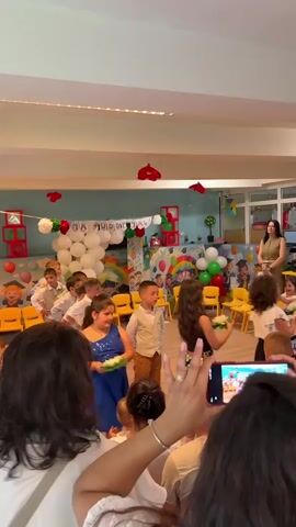 The video shows a group of young children, mostly boys, dressed in white shirts and khaki pants, standing in a line in a brightly decorated room. They are participating in some kind of performance or graduation event, with a woman in a blue dress assisting a couple of them. Two girls in floral dresses are dancing in the middle of the group, each holding what looks like a green plate with something white on it.

Behind the children, there's a decorated backdrop with balloons, colorful boxes, and a banner that says "НА ДОБЪР ПЪТ!" in Bulgarian, which translates to "Good Luck!" or "Bon Voyage!".  Yellow chairs are scattered around the room, some arranged in a semicircle.  A woman in a beige dress is standing off to the side, likely a teacher or event organizer.

In the foreground, a group of adults, presumably parents or family members, are watching and recording the event with their phones. One woman in particular is prominently holding up her phone to record. The room is well-lit with a light-colored floor.