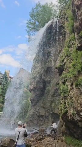 The video shows a tall, rocky cliff face with a waterfall cascading down it. The water sprays out widely at the bottom. Two people are visible at the base of the waterfall. On the left, a person wearing a grey hoodie and blue shorts stands facing the waterfall, holding a phone up to take a picture. On the right, a person wearing a white long-sleeved shirt and dark pants stands with their back to the camera, also facing the waterfall. The ground is covered in rocks and debris. Green foliage is growing on the cliff face and on trees to the left of the waterfall. A building with a tall, pointed structure is partially visible behind the trees on the left. The sky is blue with scattered white clouds.