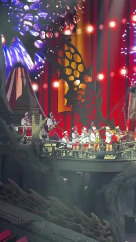 A stage performance unfolds with a brass band perched on a structure resembling a rocky bridge, complete with decorative tree-like elements. The band members are dressed in Santa Claus attire and are playing various instruments. Behind them, a backdrop of red and white curtains is illuminated by stage lights and pyrotechnic sparks. The camera then pans down to reveal a group of performers on motorcycles riding onto the stage, waving checkered flags. They are accompanied by a group of dancers. The stage is bathed in bright spotlights.