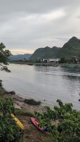 The video shows a wide river with several boats docked and moving on it. On the left bank, there are kayaks and other watercraft. In the background, a line of houses is visible along the river, with lush green, rocky hills rising behind them. The sky is overcast, suggesting a cloudy or potentially rainy day. The water appears calm, with gentle ripples. The overall scene depicts a peaceful, natural landscape with human activity centered around the river.