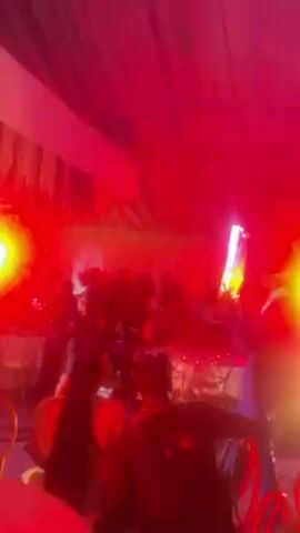 The video shows a crowded indoor event with a group of people dancing. The room is lit with bright red and blue lights, creating a vibrant atmosphere. Some individuals are holding glowing light sticks, adding to the visual spectacle. There are tables and chairs visible in the background, suggesting a party or celebration. The ceiling is draped with fabric, contributing to the overall festive setting. The camera is held by someone moving through the crowd, providing a dynamic perspective of the event.