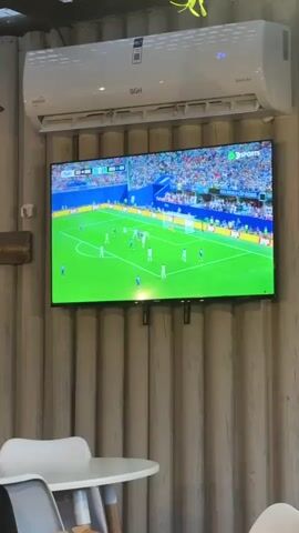 A wall-mounted flat-screen television displays a soccer match in progress, with players on a green field and a crowd in the background. Above the television, a white air conditioning unit is mounted. The television is attached to a wall with a corrugated texture. In the foreground, a white table and chairs are visible. The video shows a brief sequence of the soccer game, including a goalkeeper lying on the ground with the ball.
