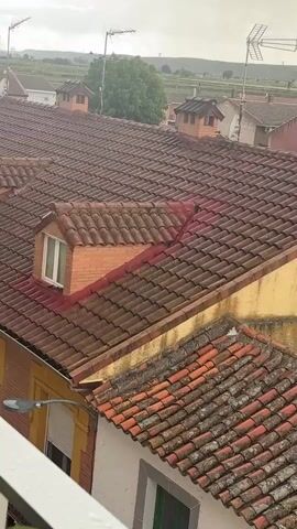From an elevated position, the camera records a cityscape with a focus on tiled rooftops under a cloudy sky. The roofs are predominantly brown and orange, showing signs of age and weathering. Dormer windows with white frames are integrated into some of the roofs. Antennas are visible atop several buildings. In the background, a green field stretches towards the horizon under the overcast sky. Raindrops are visible on the camera lens and on the window sill in the foreground.
