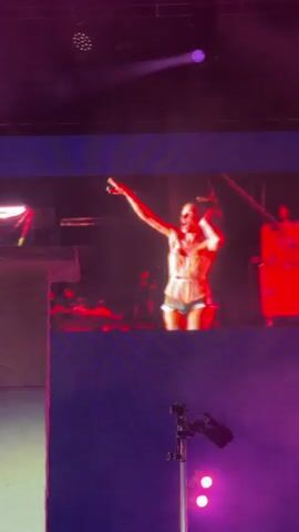 The video displays a screen showing a woman with long, curly hair, wearing a light-colored garment. She is on a stage, illuminated by red and purple lights, with her arms outstretched and her head tilted back. The screen is positioned above a stage with various equipment, including lighting rigs and microphones. The overall atmosphere appears to be a live performance or concert.