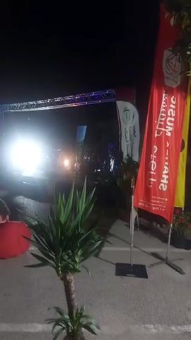 Here is a description of the video:

The scene captures a nighttime outdoor event, likely a concert or performance. A stage is set up with lights shining brightly, illuminating the performers who are visible in silhouette in the background. There's a metal truss structure over the stage. To the right of the frame, two banners stand tall. One banner is red and prominently displays the words "SHARDA MUSIC CLUB" in Arabic. The other banner, partially visible, is white with Arabic writing as well. In the foreground, a potted yucca plant adds a touch of greenery. To the left, a red beanbag is present, possibly for seating. The ground appears to be paved, and the overall atmosphere suggests anticipation and excitement surrounding the upcoming performance.