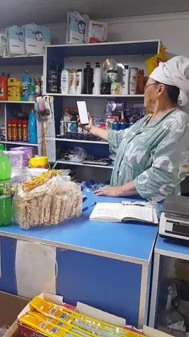 A middle-aged woman wearing a white headscarf and a light blue patterned robe stands behind a blue and white counter. She is holding a white smartphone in her right hand, angled upwards as if taking a photo or video. The phone's screen is visible and appears to display a white interface with a black circle. The woman's left hand rests on the counter. In front of her on the counter are an open book, a package of crackers, and a green bottle. To her right is a black digital scale. Behind her are shelves stocked with various products, including bottles of shampoo and cleaning supplies, as well as some boxes and bags. To the left of the counter, a box filled with yellow and orange packaged items is visible. The environment appears to be a small shop or convenience store.