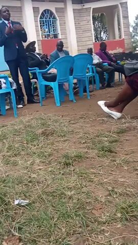 The video shows a group of people seated in blue plastic chairs in front of a brick building. The people are dressed in a variety of clothing, including suits and casual wear. One man stands and speaks. The ground is dirt and grass. A woman in white shoes walks by the seated people.