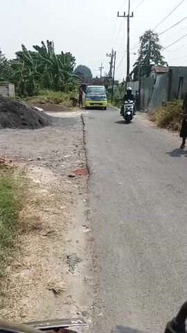 A motorcycle is driving down a paved road. To the left of the road is a dirt path with a pile of dark material, possibly soil or sand, and a stack of white building blocks. Further down the road, a yellow truck is parked. Two people are near the truck, and another person is standing on the right side of the road. The environment appears to be rural or a developing area, with vegetation visible behind the pile of material and a utility pole alongside the road. The sky is clear and bright, suggesting it is daytime.