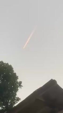 The video shows a bright, thin orange trail in the sky, possibly from a plane or rocket, against a hazy light grey sky. In the lower left corner, there's a tree with green leaves, and in the lower right, part of a wooden roof with visible beams is visible. The camera appears to be stationary, capturing the sky and these foreground elements.
