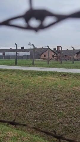The video shows a view of brick buildings with rows of chimneys, surrounded by barbed wire fences. The buildings appear to be old and industrial. There is a grassy area in the foreground and a road or path in the middle ground. In the distance, a few people can be seen walking near the buildings. The sky is overcast. The overall impression is one of a somber and desolate place.