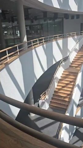 This is a video of the interior of a modern, multi-story building. The scene is dominated by a grand, curved staircase made of wood and metal railings, leading upwards through several levels. Sunlight streams in from the ceiling, casting dramatic shadows across the white walls and staircases, suggesting it is daytime. The architecture is sleek and contemporary, with clean lines and open spaces.

The upper floors are visible, showcasing the building's unique circular design. We see balconies and walkways overlooking the central atrium, with railings and further staircases. Some areas appear to be under construction or renovation, indicated by exposed elements and temporary barriers. On the ground floor, near the base of the main staircase, a green directional sign with a white arrow and a symbol of a person walking can be seen.

The overall impression is one of a spacious, light-filled public or institutional building, possibly a library, university building, or modern office complex. The absence of people suggests the building is either empty or this area is not currently in use. The lighting and the architectural style evoke a sense of calm and order. There is a faint red and white striped tape visible across the stairs in the middle ground, possibly a caution tape. The time of day appears to be during daylight hours, judging by the strong sunlight.