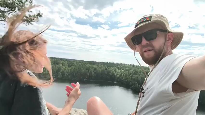 Two people, a woman with long brown hair and a man wearing a bucket hat and sunglasses, are sitting on a rocky outcrop overlooking a lake. The woman is in the foreground, looking over her shoulder at the camera with a smile. The man is to her right, facing the camera and speaking. Behind them, a dense forest surrounds the lake, and the sky is partly cloudy. The man is holding a phone, presumably recording the video. There are backpacks and other gear visible in the foreground. The overall scene is a scenic outdoor setting.