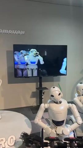 The video shows a television screen displaying a white robot playing a trumpet. The robot on the screen is wearing a blue band around its neck and is positioned in front of a dark background. In the foreground, a white robot with a Toyota logo on its chest is seated. Another white robot is partially visible to the right. The robots are in what appears to be an exhibition or museum setting, with a wall featuring the word "tomorrow" in white text.