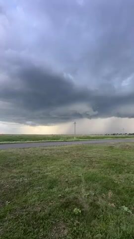 This is a vertical video clip taken outdoors in the village of Ozyorny, Kazakhstan. The scene depicts a vast, open landscape with a prominent stormy sky dominating the frame. Dark, heavy storm clouds are gathered overhead, with shafts of rain visible in the distance, creating a dramatic and somewhat ominous atmosphere.

The foreground consists of a grassy field, with a paved road cutting through the middle ground. A single utility pole stands near the road, and a few scattered structures or trees are visible in the far distance. The lighting suggests it is likely late afternoon or early evening, given the subdued light and the overcast conditions.

The primary activity captured is the approaching or ongoing rain shower associated with the storm. There are no people or specific objects of focus in the foreground, emphasizing the natural event and the expansive scenery. The overall mood evoked is one of raw, powerful nature, with the approaching storm conveying a sense of anticipation or perhaps a slight unease. There is no visible text in the video.