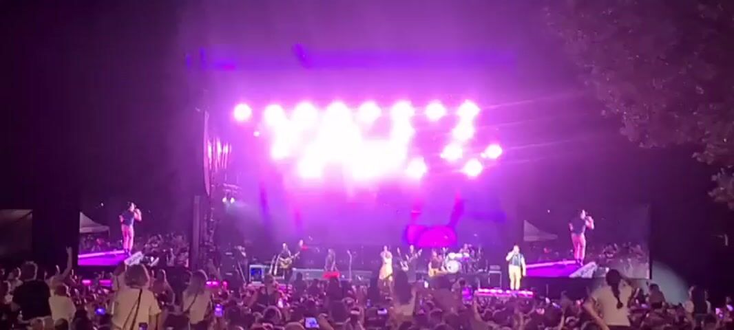 A large crowd of people are gathered in front of a stage at night. The stage is brightly lit with pink and white lights. Several musicians are performing on stage. The audience is holding up their phones to record the performance. Some people in the crowd are standing on the shoulders of others. The overall atmosphere is energetic and festive.