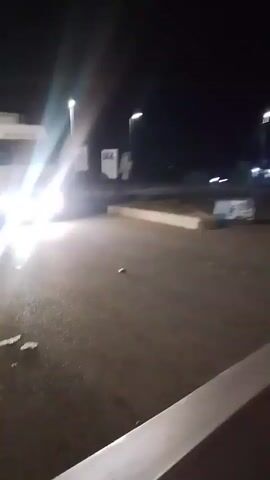 This video shows a semi-truck at night, likely in a parking lot or rest stop. The truck is white with a large trailer. Its headlights are on and very bright, obscuring details of the front of the vehicle. The ground appears to be asphalt and is wet, suggesting recent rain or damp conditions.

In the background, there are streetlights and the dark silhouettes of other vehicles and structures. The scene is dimly lit overall, with the truck's headlights being the dominant light source. There is some debris scattered on the ground in front of the truck.

The camera is positioned low and close to the ground, possibly from within another vehicle or behind an object. The motion of the camera suggests it might be mounted on a vehicle that is moving or has just stopped. The overall atmosphere is nocturnal and somewhat utilitarian, typical of a truck stop or industrial area at night. There is no visible text that can be clearly read.