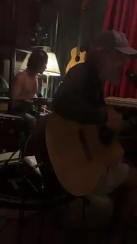 Here's a description of the video:

The video shows a man playing an acoustic guitar and singing into a microphone. He's wearing a long-sleeved dark shirt, khaki shorts, and a grey baseball cap. He has a grey beard. Behind him, to the left, a shirtless man is playing the drums. A neon sign reading "MACHINE" is visible in the upper left corner of the frame. There is a lamp and a keyboard in the background, as well as two guitars hanging on the wall. A red curtain is visible on the right side of the frame. The room appears to be a music studio or a small performance space.