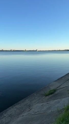 A vertical video shows a calm body of water with a clear blue sky above. In the foreground, a concrete embankment slopes down towards the water. A few blades of green grass are visible growing on the embankment. In the distance, a faint line of buildings marks the horizon. The water is dark blue and reflects the sky, with subtle ripples indicating gentle movement. The overall impression is one of tranquility and open space.