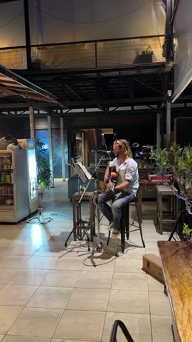 A man with long blonde hair and sunglasses is playing an acoustic guitar while sitting on a stool. He is wearing a light-colored button-down shirt and jeans. A microphone is positioned in front of him, and a music stand with papers is to his left. He is in an outdoor setting with tiled flooring. In the background, there is a refrigerator on the left, plants, and tables and chairs. The lighting suggests it is evening.