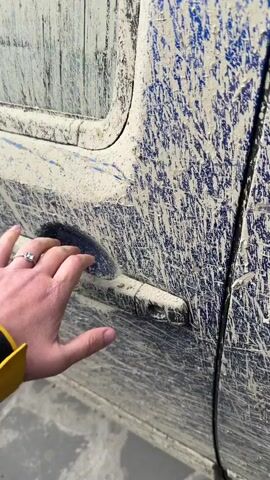A person wearing a yellow jacket opens the driver's side door of a heavily mud-covered blue vehicle. Inside the door compartment are crumpled white tissues and a bottle. The car's interior, including the floor mat and seat, also shows evidence of mud. The ground outside is concrete.