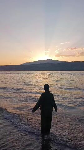A person wearing a dark hooded jacket and dark pants stands in shallow water at the edge of a body of water. The sun is setting behind a mountain range on the horizon, casting rays of light through the clouds. The water is calm with gentle waves. The person is facing away from the camera, looking towards the sunset. The person turns their head to the left and walks forward.