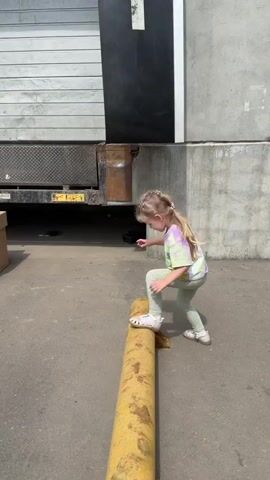 A young girl with blonde hair in pigtails, wearing a tie-dye shirt, polka-dotted leggings, and white sandals, is attempting to balance on a long, yellow, cylindrical bollard. The bollard is situated on a grey concrete surface in front of a loading dock. The dock has a partially open door showing a metal loading platform. The girl is looking down at her feet as she tries to keep her balance. The environment appears to be an industrial or commercial area.