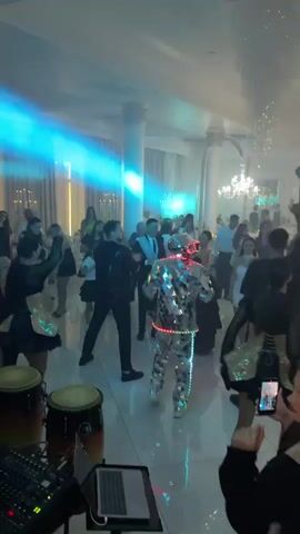 A group of people are dancing in a brightly lit room. In the center of the room, a person in a mirrored, illuminated costume is dancing. Several people are filming the person in the mirrored costume on their phones. In the foreground, there are two conga drums and a DJ mixer. The room is decorated with chandeliers and white curtains.