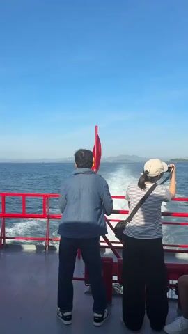 A static, eye-level shot shows the rear view of two people standing on the deck of a ferry. The person on the left, wearing a denim jacket and dark pants, faces away from the camera and towards the water. A red Vietnamese flag is prominently displayed behind them. To the right, a person in a striped shirt and black pants, wearing a light-colored baseball cap, holds a phone up, seemingly taking a photo or video. Both individuals are looking out at the ocean, which stretches to the horizon under a clear blue sky. The ferry's wake trails behind it, creating a foamy white path in the water. Red railings with white accents line the edge of the deck. The overall atmosphere is peaceful, suggesting a scenic journey.