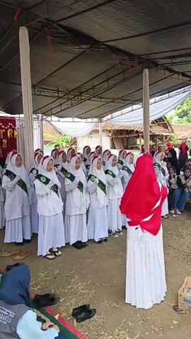 A group of young girls in white hijabs and matching white uniforms with green sashes are standing in rows under a tent, seemingly singing or performing. Their hands are held together in front of them. One girl, who is taller and appears to be a leader or instructor, stands facing the group, wearing a red hijab and a white dress. She is positioned slightly to the right of the group of girls. In the background, there are more people seated, some in the shade of the tent and others outside. The environment appears to be outdoors, with a dirt ground and some scattered hay or straw. There are white poles supporting the tent, and the overall setting suggests some kind of outdoor gathering or event.