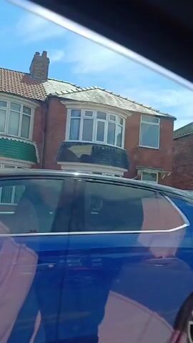 This media file captures a drive through a residential street in Redcar, United Kingdom. The view is from inside a vehicle, likely through the windshield or side window, with the edge of the vehicle's interior visible in the foreground.

The scene primarily features terraced houses made of red brick, typical of many UK residential areas. These homes have bay windows, pitched roofs, and front gardens with fences and some greenery. Several cars are parked on driveways or on the street, including a light blue Fiat 500, a blue Nissan, and a black car. One shot also shows a red car passing by. The houses appear to be well-maintained.

The activity is a slow drive or journey along the street. The time of day is clearly daytime, with bright sunlight casting shadows. The weather appears to be pleasant, with a blue sky and some clouds visible. The presence of a license plate on the blue Fiat 500 (visible as "NDV" followed by other characters) and the style of the houses and cars suggest the United Kingdom as the location. No people are clearly visible in the external shots, though the interior view hints at the presence of passengers. No specific emotions are conveyed, and there is no discernible text other than the partial license plate.