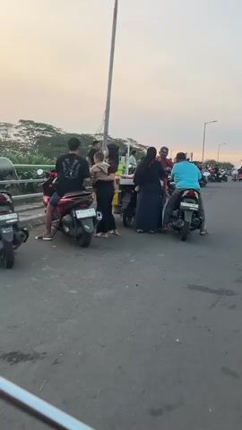 The video captures a street scene in Besuki, Indonesia, likely during the late afternoon or early evening given the soft, warm light and the long shadows. The setting appears to be a road or pathway alongside an open area, possibly a park or a waterfront, with a few palm trees visible in the distance.

Several people are present. In the foreground, a woman wearing a black dress and carrying a baby on her hip walks away from a stall. Next to her, another woman in a dark hijab and long blue skirt is interacting with a man in a red shirt and blue cap, who appears to be a vendor at a colorful stall with a green and yellow structure. Behind them, a man wearing a bright blue t-shirt is seated on a black motorcycle, facing away from the camera. More motorcycles are parked or moving in the background, suggesting a busy public space. The overall atmosphere seems relaxed and communal, with people engaged in casual social interaction and likely enjoying the cooler part of the day.

The weather appears to be clear and pleasant, with a soft, overcast sky suggesting sunset is approaching. The road surface is asphalt. There is a tall, thin metal pole to the left of the frame, possibly a lamppost or part of a structure.

Visible text includes the license plate on the motorcycle in the foreground, which reads "W 5529 NGI".