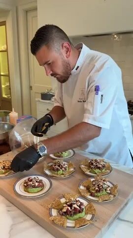 A male chef with short, dark hair and a beard, wearing a white chef's coat with red and green trim and black gloves, is carefully garnishing small plates of food. Each plate contains a circular serving of what appears to be a tuna or salmon tartare mixture on top of mashed avocado, with a drizzle of sauce and a few tortilla chips arranged around the base. The chef is using tweezers or small tongs to place a garnish, possibly a sprig of herbs or a tiny edible flower, onto one of the dishes. He is positioned behind a wooden cutting board that holds at least eight of these prepared plates, arranged in two rows. To the left of the chef, a metal bowl and a squeeze bottle with a red sauce are visible. The environment appears to be a well-lit kitchen with a marble countertop and cabinetry in the background. The chef's focus is entirely on the precise placement of the garnish.