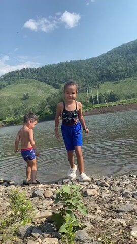 This media file captures a candid moment of two children by a river on a sunny day in Maloarslangulovo, Russia. The foreground shows a rocky riverbank with sparse green vegetation. In the shallow water, a young boy is crouched down, seemingly playing with pebbles. He is wearing blue and red swimming shorts. Further back and closer to the camera, a young girl stands on the bank, facing the water. She is wearing a black swimsuit with a colorful print and blue shorts, along with white sneakers. Her pose suggests she is either about to enter the water or is interacting with the person filming.

The background features a wide, calm river flowing between the rocky bank and a verdant, hilly landscape. The hills are covered in dense green trees, indicating a summer or early autumn setting. The sky is a clear blue with a few scattered white clouds. The presence of power lines running across the landscape suggests proximity to a populated area, even in this natural setting.

The overall mood is playful and carefree, typical of children enjoying outdoor activities. The sunlight indicates it's daytime, likely mid-morning or afternoon. The clear sky and green landscape point to pleasant weather conditions. The camera angle is low, making the children appear prominent against the backdrop. At one point, the girl's hands appear in the frame, making a peace sign, and then her face leans into the frame, suggesting she is aware of being filmed and is engaging with the camera. No visible text is present in the media file.