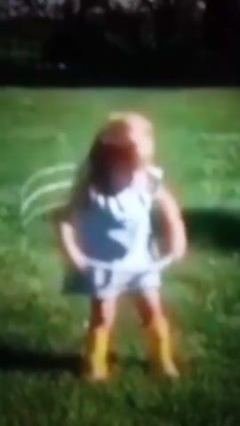 A blurry video shows a young child with blonde hair, wearing a white dress and yellow boots, dancing outdoors on a grassy lawn. The child is spinning and appears to be holding a thin, horizontal object. The background is out of focus, with hints of trees and shadows suggesting a sunny day. The video is very low resolution and has a grainy quality.
