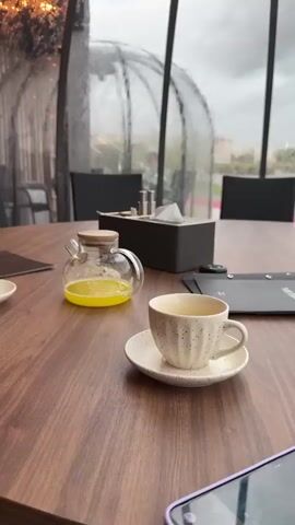 The camera is situated inside a transparent, dome-like structure with a black metal frame, showing a wooden table set for dining. On the table, there's a cup and saucer, a glass teapot filled with a yellow liquid, a black rectangular box containing napkins and possibly condiments, and a black menu. The background shows a view of a landscape obscured by heavy rain on the dome's exterior. Outside, buildings and green vegetation are visible through the rain-streaked glass. The weather appears overcast and stormy. The camera focuses on the rain pouring down outside, obscuring the view. There are black chairs surrounding the table, but they are unoccupied, and the scene appears quiet and still.