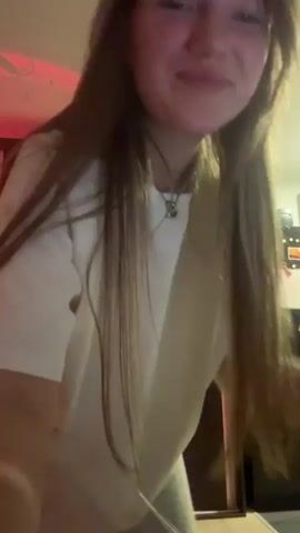 A young woman with long brown hair dances in a brightly lit room with red accents. She wears a loose, light-colored T-shirt and matching bottoms. A man suddenly appears behind her, leaning in and dancing. He's fair-skinned with short hair and wears a black shirt. The camera angle shifts and the background shows a white wall with black shelves, a red refrigerator, and a small white object. The lighting is bright, and the camera moves erratically due to the dancing.