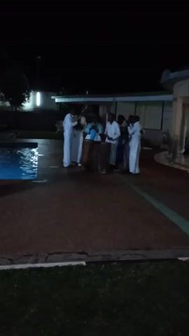 The video takes place at night on a patio next to a swimming pool. There are about six people dressed in long white robes standing in a group. Then, two women come forward. They are wearing pink skirts and white draped wraps. They perform a synchronized dance, moving their arms out to the sides and turning. There are trees and buildings in the background. The area is dimly lit, with the pool illuminated by a blue light.