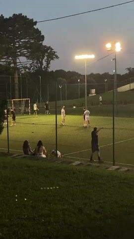 A group of people are playing soccer on a lit field at dusk. The field is surrounded by a fence and grass. Several people are watching the game from the sidelines. The sky is dark, indicating nighttime. The lighting on the field is bright, illuminating the players and the game.