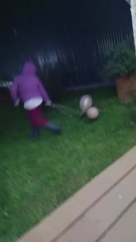 A person in a purple jacket and dark pants runs across a grassy lawn at dusk. They are holding a chain attached to two pale pink balloons. The person is moving from left to right across the frame. In the background, a house with lights on is visible, along with trees.