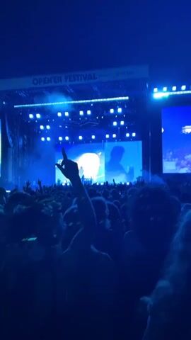 A nighttime outdoor concert is taking place. The stage is illuminated with bright blue lights and a large screen displays abstract visuals and silhouettes of performers. A crowd of people is visible in the foreground, with many of them raising their hands in the air, some holding up cell phones. The atmosphere appears energetic and celebratory. The text "OPEN'ER FESTIVAL" and "Powered by Orange" is visible at the top of the stage structure.