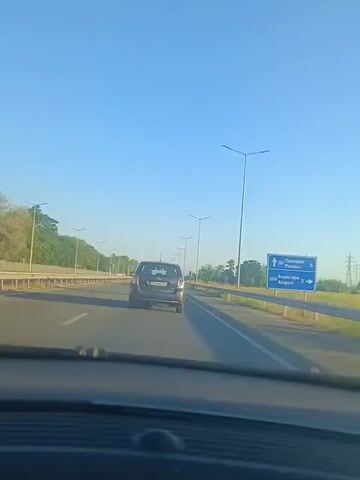 The video shows a view from inside a car driving on a highway. The camera is looking forward through the windshield. A dark colored car is in front, in the same lane. The highway has multiple lanes in each direction, separated by a median with streetlights. The left side of the highway is lined with trees and foliage. The right side has a guardrail and some grassy areas. There is a blue highway sign on the right side of the road with text in a foreign language and the word "Airport." The sky is clear and blue, indicating it is daytime. The lighting suggests it could be morning or late afternoon. The car in front has a license plate that is partially visible. There are no other people or vehicles clearly visible in the immediate vicinity.