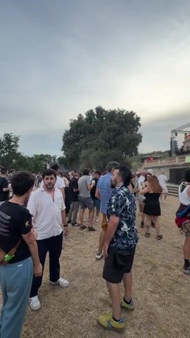 A group of people are gathered outdoors in front of a large, ornate building with a dome. There is a stage set up in front of the building with lighting equipment. Many of the people are casually dressed and appear to be enjoying themselves. Some are standing and talking, while others are dancing. The ground is covered in dry grass and dirt. The sky is overcast.