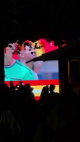 A group of people watch a large screen displaying a soccer player. The screen is adorned with the Spanish flag colors, red and yellow, and a score of "1" is visible in the upper right corner. The crowd is silhouetted against the bright screen. The ambient lighting is dim, suggesting it is nighttime. A tree branch with leaves hangs over the top of the screen.