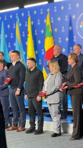 A group of men and women are gathered indoors on a raised platform, posing for a photograph. Ukrainian President Volodymyr Zelenskyy is visible in the center, dressed in a black jacket and black pants. To his left, several men wear suits or casual tops, while a woman in a gray suit stands to his right. Many individuals are holding rectangular red items, possibly awards or certificates, while some men further to the right hold blue plaques or cases.

Behind the group, several flags are displayed: multiple Ukrainian flags (blue and yellow), several yellow flags with distinct symbols (one with a starburst, another with a green leaf), and a red, yellow, and green flag, which appears to be the flag of Moldova. The background is a blue wall adorned with a repeated pattern of light blue squares containing a cross symbol. Text on the wall, partially visible, reads "КОНГРЕС МІСЦЕВОЇ," suggesting a "Congress of Local Authorities" or similar event. The overall atmosphere appears formal and celebratory, with several individuals smiling. The scene is well-lit, indicative of an official event.