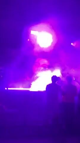 The video starts with a shot of a large screen displaying a person's face, illuminated in red and purple light. The camera then pans out to reveal a crowd of people in a dark environment with purple lighting. People are walking around, some are dancing. The camera continues to pan, showcasing the outdoor event, with canopies decorated with red fabric.
