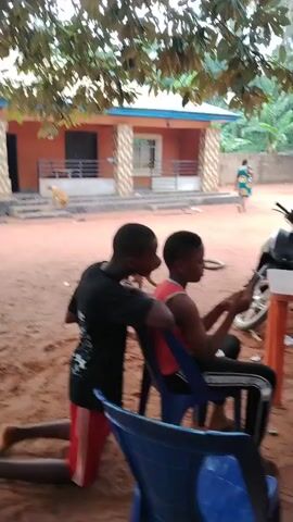 The video captures an outdoor scene, likely in Akwa Etiti, Nigeria, during the daytime. In the foreground, two young boys are focused on a smartphone. One boy, dressed in a black t-shirt and red and white striped shorts, is kneeling on the reddish-brown dirt ground, leaning over the shoulder of another boy. The second boy, wearing a red tank top and dark pants, is seated in a blue plastic chair, holding and looking at the smartphone. They appear to be sharing the device, possibly playing a game or watching something together.

Behind the boys, a small orange table holds a plastic bottle of water and some other items, with a black motorbike parked nearby. A woman in a dark outfit stands near the table, while a man is seated in a blue plastic chair further back. In the background, a light-colored building with a dark roof and pillars is visible. The scene is shaded by large green trees, and the ground is consistently reddish-brown dirt. The overall atmosphere is casual and relaxed, suggesting a common social or leisure activity in an outdoor setting.