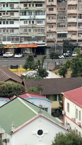 The video shows an elevated view of a street scene with several buildings. In the foreground, there are rooftops of houses, some with green and red corrugated metal roofs. Behind these houses, a multi-story apartment building dominates the background. This building appears to be made of concrete and has many windows and balconies. The street level has shops with awnings and parked cars. Trees are scattered throughout the scene, some growing between the lower houses and the apartment building. The sky is overcast, suggesting a cloudy day. There are no visible people or ongoing actions that can be specifically identified or described beyond their presence.