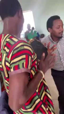 Here's a description of the video:

The video shows a group of people in a room, possibly a school setting. A person in a colorful striped shirt plays a handheld drum, sings, and dances. An adult male in a patterned button-down shirt and dark pants dances alongside them. Other individuals, presumably students in uniforms, are visible in the background dancing and moving around. The room has light green walls and a tiled floor.  The lighting appears to be from a window or overhead fixtures. The focus is primarily on the drumming and dancing happening in the foreground.