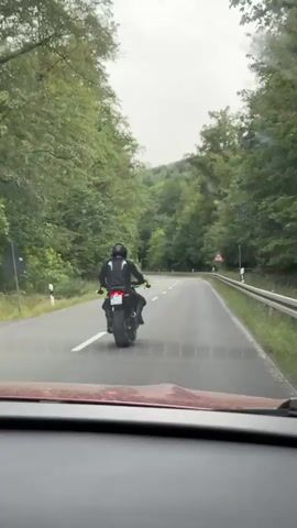 A person wearing a black motorcycle suit and helmet rides a black motorcycle on a paved road. The road is surrounded by a dense forest with green trees. The motorcycle has a visible license plate. The rider is in the process of making a left turn. A road sign indicating a curve to the right is visible on the right side of the road. The video appears to be recorded from another vehicle following the motorcycle.