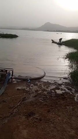 The scene shows a dam with a man standing next to a small boat at the edge of the water. The water is still and reflects the sunlight. A pump and hose are located on the muddy bank next to the water, connected to the water source. The sky is overcast. A mountain is visible in the background. Another boat is further out in the water. There are patches of green grass along the shoreline. The shore consists of muddy terrain. A tire, a bucket, and a plastic container are on the shore next to the pump.